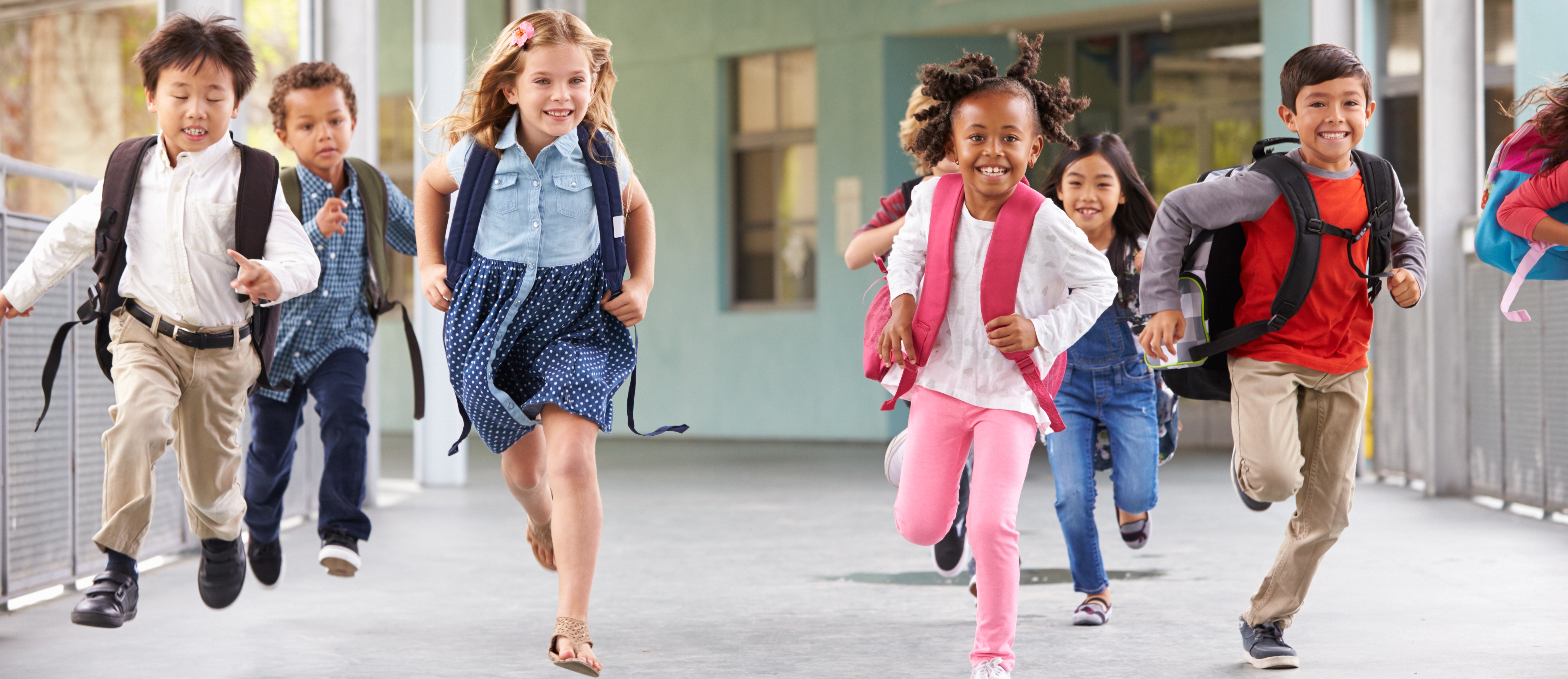 A diverse group of young children, wearing backpacks and smiling as they run across a school walkway